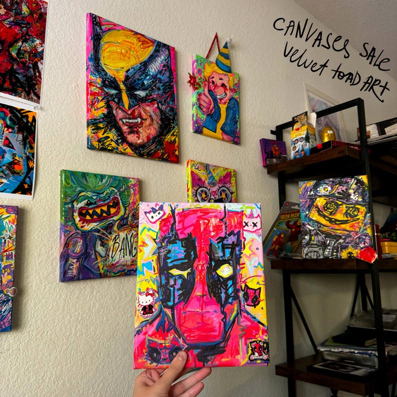 May include: A collection of colorful, abstract paintings on canvas. The paintings feature various characters and designs, including a pink and black Deadpool, a green and yellow monster, a yellow and red Wolverine, and a yellow and red character with a party hat. The text 'CANVASES SALE velvet toad ART' is written in black marker on a white background.