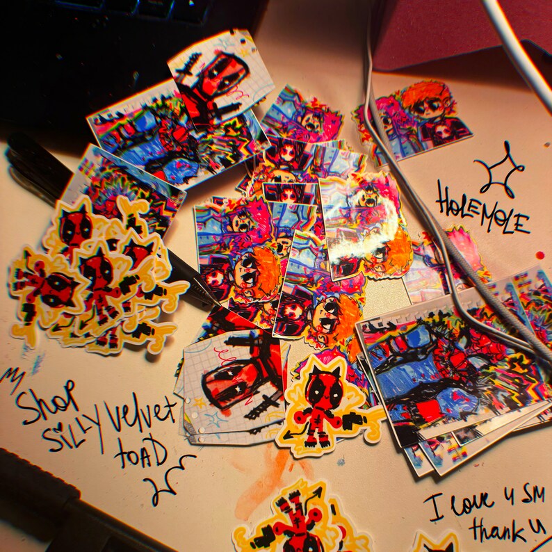 May include: A collection of colourful stickers featuring cartoon characters, including Deadpool and a cat-like figure. The stickers are on a white surface with handwritten text that reads "Shop Silly Velvet Toad".