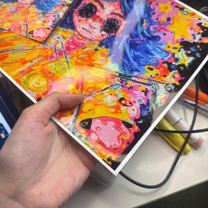 May include: A colorful art print featuring a whimsical illustration of a character with blue hair and large eyes. The artwork is vibrant with shades of yellow, pink, orange, and blue. The print is held by a hand, showcasing the glossy finish and detailed design.