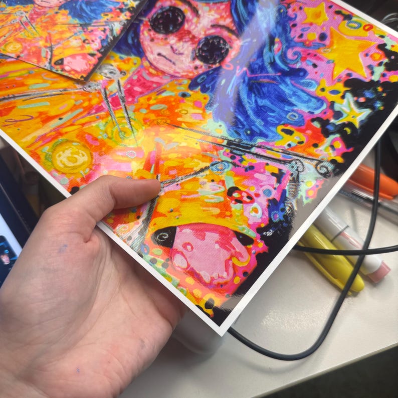 May include: A colorful art print featuring a whimsical illustration of a character with blue hair and large eyes. The artwork is vibrant with shades of yellow, pink, orange, and blue. The print is held by a hand, showcasing the glossy finish and detailed design.