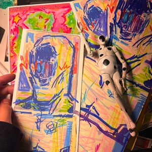 Pu&ograve; includere: Una action figure bianca con pois neri &egrave; sdraiata su una pila di fogli colorati con disegni astratti. I fogli sono coperti di colori vivaci, tra cui rosa, blu, verde e giallo.