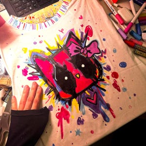 May include: A white t-shirt with a hand-drawn cat face design in bold colours. The design features a pink bow, black, red, yellow, and blue accents, and paint splatters. The collar has a colourful striped pattern. The overall style is artistic.