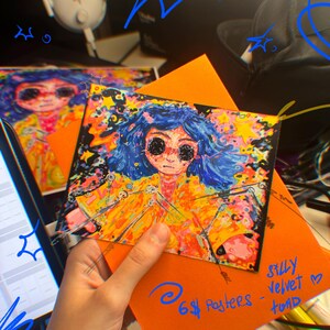 May include: A colorful art print featuring a character with blue hair and black eyes, wearing a yellow coat. The print is held over an orange envelope. Text on the envelope reads "6$ Posters - Silly Velvet - toad."