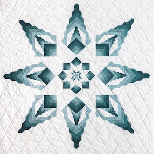 Pode incluir: Um edredom branco com um grande desenho de floco de neve azul-esverdeado no centro. O floco de neve é feito de várias camadas de formas geométricas, criando um efeito 3D.