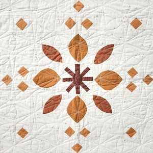 May include: A white quilt with a central floral design. The design features brown and orange leaf shapes and diamond accents. The quilt is stitched with a geometric pattern and small gray star-like details.