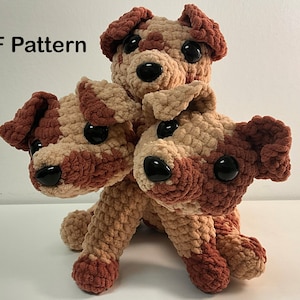 May include: A three-headed crochet dog in brown and beige tones. The soft toy has large, black button eyes and is made from a textured yarn. The words "PDF Pattern" are visible in the upper left corner.
