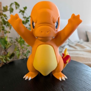 May include: Orange Charmander figurine with arms raised, featuring a yellow belly and a flame-shaped tail. The figure has blue eyes and white claws. The figurine is a collectible item.