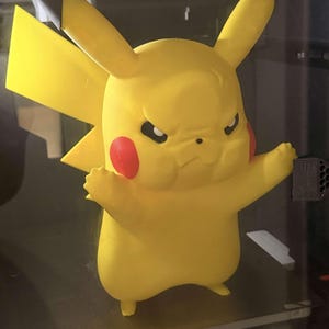 May include: A yellow Pikachu figurine with a stern expression, red cheek circles, and black-tipped ears. The figure is holding a small, dark gray object. The figurine is a popular character from the Pokémon franchise.