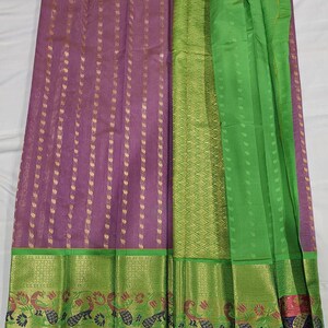 May include: A vibrant textile featuring a purple body with vertical gold accents. The design includes a green border with floral and peacock motifs. The fabric is partially draped, showcasing its texture and color variations.
