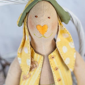 May include: A handmade plush bunny toy with a yellow and white striped vest, a green hat, and a heart-shaped nose. The bunny has brown eyes and a stitched smile.