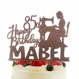 A birthday cake with a glittery topper that says "Happy Birthday Mabel" and features a silhouette of a woman sewing.