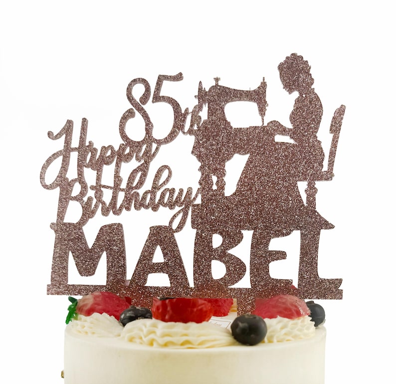 A birthday cake with a glittery topper that says "Happy Birthday Mabel" and features a silhouette of a woman sewing.
