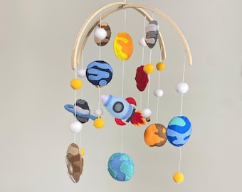 May include: A wooden mobile with felt planets, stars, and a rocket ship. The planets are blue, green, orange, and red. The stars are yellow. The rocket ship is blue and red.