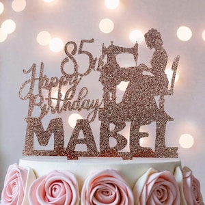 A birthday cake with a glittery topper that says "Happy Birthday Mabel" and features a silhouette of a woman sewing.
