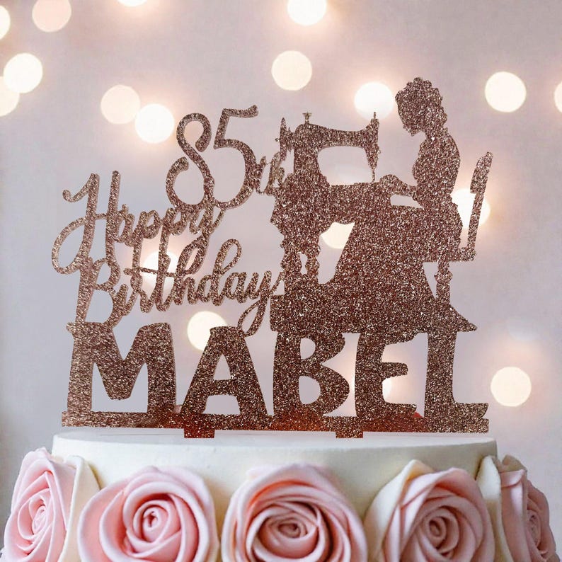 A birthday cake with a glittery topper that says "Happy Birthday Mabel" and features a silhouette of a woman sewing.