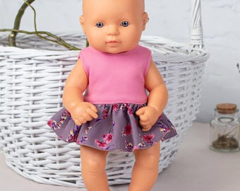 May include: A baby doll with a smooth peach-toned complexion and blue eyes, wearing a sleeveless top with a solid pink bodice and a flared skirt featuring a dark grey base with a vibrant pink and purple floral pattern printed throughout the fabric.