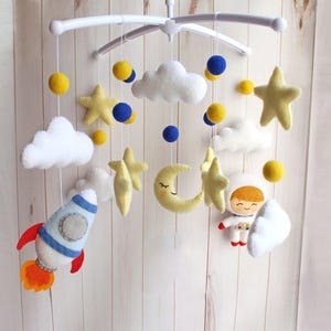 Puede incluir: Móvil para bebé hecho a mano con estrellas, nubes, lunas y un astronauta de fieltro. El móvil incluye un cohete y detalles en amarillo y azul. Decoración de guardería suspendida de un marco de plástico blanco.