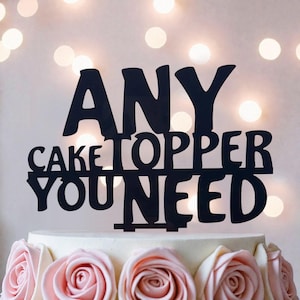 May include: A black cake topper featuring the text ANY CAKE TOPPER YOU NEED in a bold, sans-serif font. The topper is positioned on top of a white cake decorated with a row of light pink frosting roses arranged along the bottom edge.