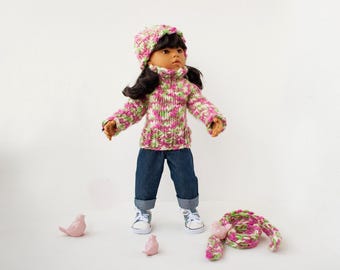 Può includere: Una bambola con un maglione di lana rosa e verde, un cappello abbinato e jeans blu. La bambola è in piedi su una superficie bianca con uccelli di ceramica rosa e una sciarpa di lana rosa e verde.