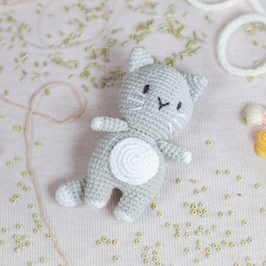 May include: A gray crocheted cat toy with white accents. The cat has a white circle on its belly and black eyes and whiskers.