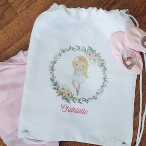 May include: White drawstring bag with a floral wreath design featuring a ballerina girl. The bag has the name "Charlotte" printed below the design. Two pink ballet shoes are lying on top of the bag.