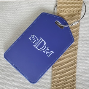 May include: A blue luggage tag with the initials "SDM" in white lettering. The tag is rectangular with rounded corners and is attached to a silver-colored wire loop. The tag is resting on a beige canvas bag.
