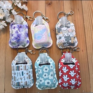 May include: Six colorful hand sanitizer holders with keychains. The holders are made of fabric and have different patterns, including floral, tie-dye, cats, books, flowers, and Mickey Mouse hands.