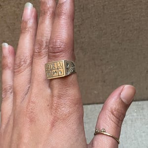 Puede incluir: Un anillo de plata con una parte superior cuadrada de oro con dise&ntilde;os intrincados. El anillo se lleva en el dedo &iacute;ndice de una mano.