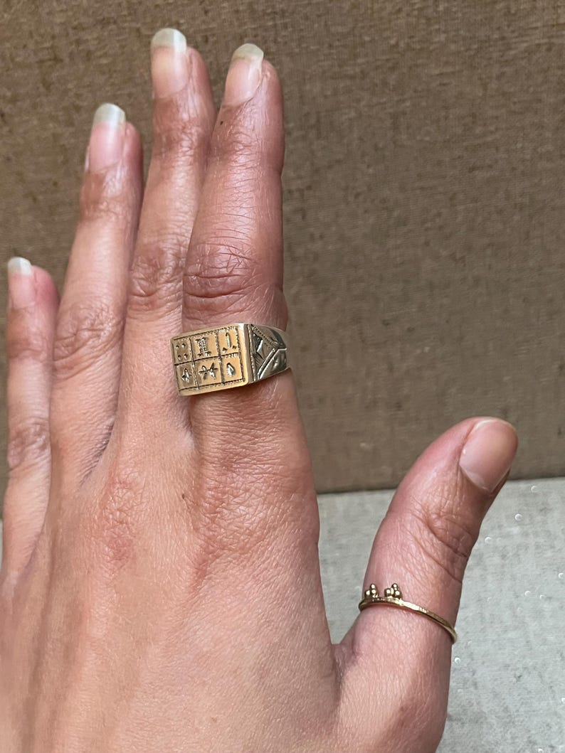 Puede incluir: Un anillo de plata con una parte superior cuadrada de oro con dise&ntilde;os intrincados. El anillo se lleva en el dedo &iacute;ndice de una mano.