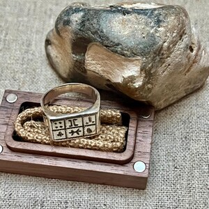 Puede incluir: Un anillo de plata con una cara cuadrada que presenta un dise&ntilde;o de varios s&iacute;mbolos. El anillo se muestra en una caja de madera con una cuerda marr&oacute;n.