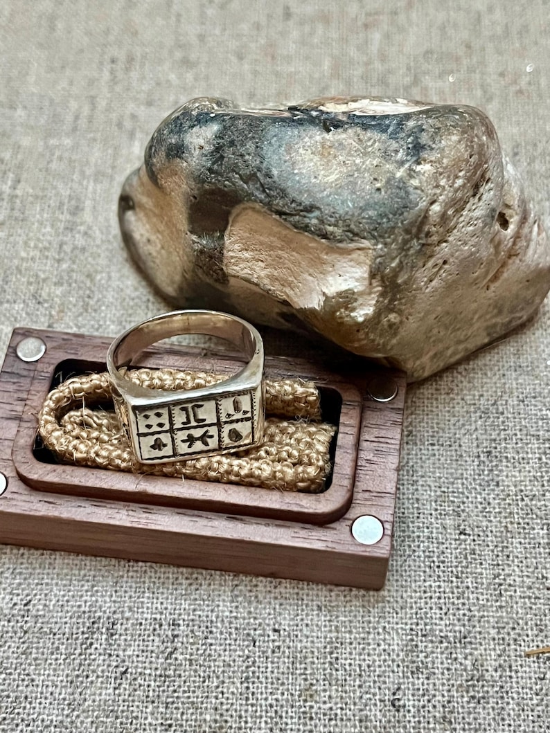 Puede incluir: Un anillo de plata con una cara cuadrada que presenta un dise&ntilde;o de varios s&iacute;mbolos. El anillo se muestra en una caja de madera con una cuerda marr&oacute;n.