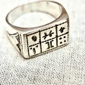 Puede incluir: Un anillo de plata con un dise&ntilde;o cuadrado que presenta un patr&oacute;n de s&iacute;mbolos y puntos.