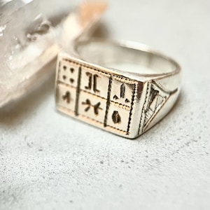 Puede incluir: Un anillo de plata con un dise&ntilde;o cuadrado que presenta grabados intrincados. El anillo tiene un acabado pulido y una banda ligeramente redondeada.