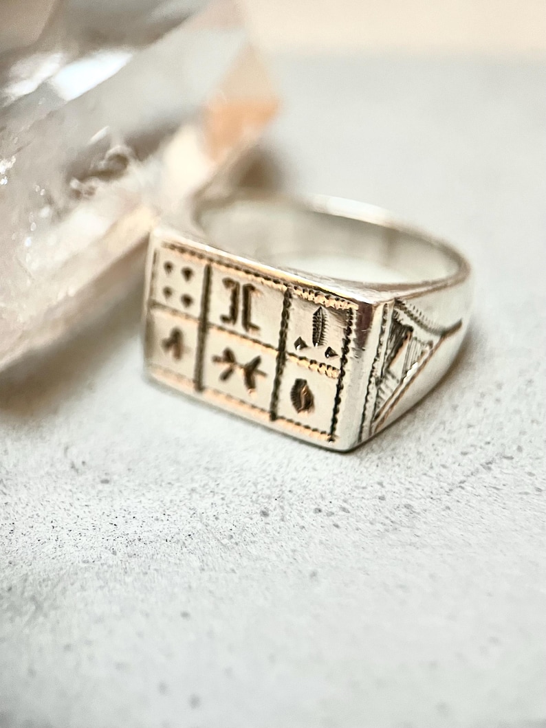 Puede incluir: Un anillo de plata con un dise&ntilde;o cuadrado que presenta grabados intrincados. El anillo tiene un acabado pulido y una banda ligeramente redondeada.