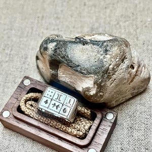 Puede incluir: Un anillo de plata con un dise&ntilde;o cuadrado que presenta varios s&iacute;mbolos y un borde texturizado. El anillo se muestra en una caja de madera con una cuerda marr&oacute;n.