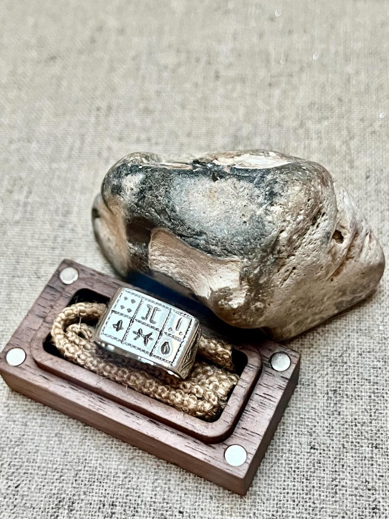 Puede incluir: Un anillo de plata con un dise&ntilde;o cuadrado que presenta varios s&iacute;mbolos y un borde texturizado. El anillo se muestra en una caja de madera con una cuerda marr&oacute;n.