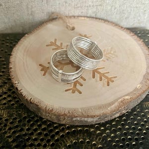 Puede incluir: Dos anillos de plata con un diseño texturizado, apilados uno encima del otro sobre una superficie de madera con un diseño de copo de nieve.