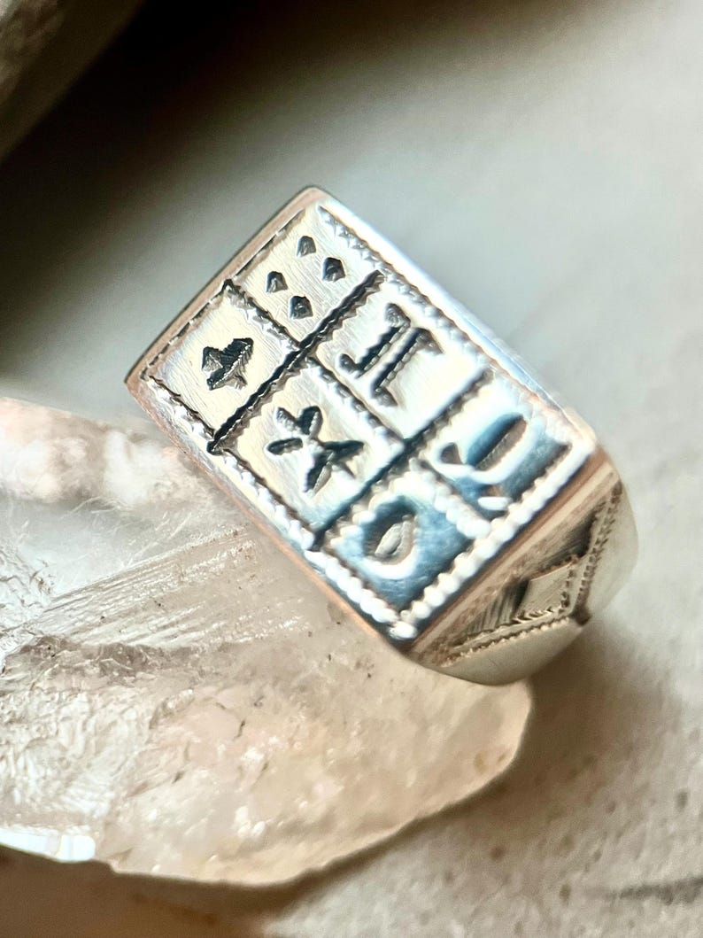 Puede incluir: Un anillo de plata con una cara cuadrada que presenta un dise&ntilde;o intrincado de cuatro cuadrados con diferentes s&iacute;mbolos en cada uno. El anillo tiene un borde texturizado.