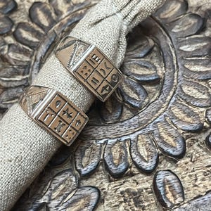 Puede incluir: Dos anillos de plata con dise&ntilde;os intrincados, con forma cuadrada y s&iacute;mbolos, se muestran sobre una superficie marr&oacute;n texturizada. Los anillos est&aacute;n anidados en una peque&ntilde;a bolsa de tela beige.