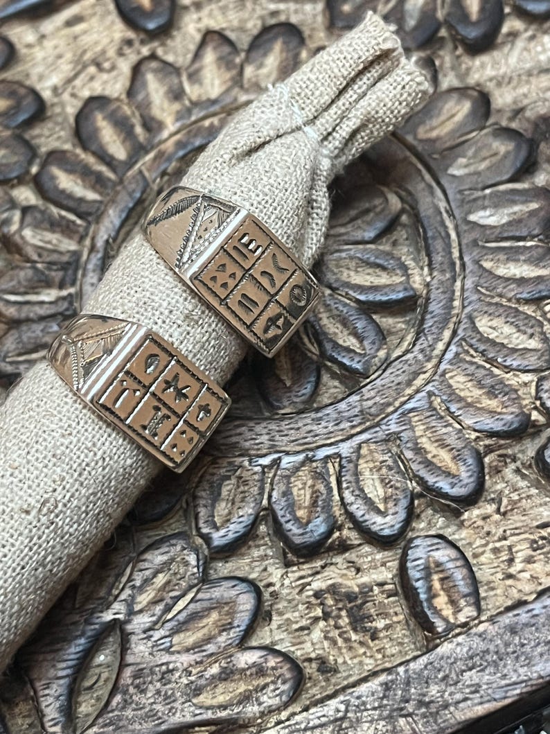 Puede incluir: Dos anillos de plata con dise&ntilde;os intrincados, con forma cuadrada y s&iacute;mbolos, se muestran sobre una superficie marr&oacute;n texturizada. Los anillos est&aacute;n anidados en una peque&ntilde;a bolsa de tela beige.