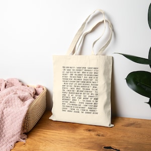 Peut inclure: Un sac fourre-tout de couleur crème avec du texte noir. Le texte comprend des phrases comme « Tu es jolie » et « Je pourrais ouvrir ». Le sac a un design simple et est probablement en toile ou dans un matériau similaire. Le sac est à côté d'un panier tressé.
