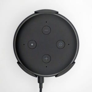 May include: A black smart speaker with a circular design. The top surface features volume control buttons, a microphone mute button, and an action button. A black power cord extends from the bottom.