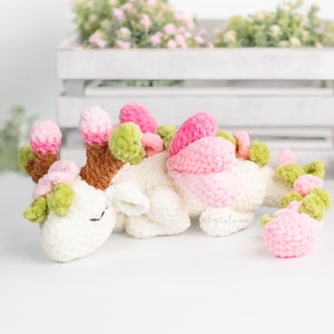 May include: A white crocheted dragon with pink and green flowers on its back, lying on a white surface.