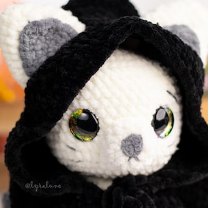 May include: A white crocheted cat wearing a black hooded cloak. The cat has large, sparkly eyes and a grey nose.