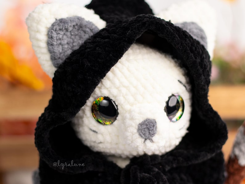 May include: A white crocheted cat wearing a black hooded cloak. The cat has large, sparkly eyes and a grey nose.