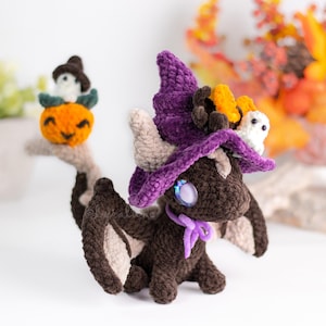 Puede incluir: Un dragón marrón tejido a ganchillo con un sombrero de bruja morado, un lazo lavanda y ojos azules grandes. Una pequeña calabaza con una cara sonriente y un pequeño sombrero de bruja se encuentra en una rama. Un fantasma blanco está posado en el sombrero del dragón.