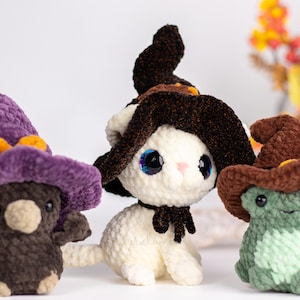 May include: Three handmade crochet plushies: a brown bear, a white cat, and a green frog, each wearing a witch's hat. The bear has a purple hat with orange accents. The cat has a black bow. The frog has a brown hat.