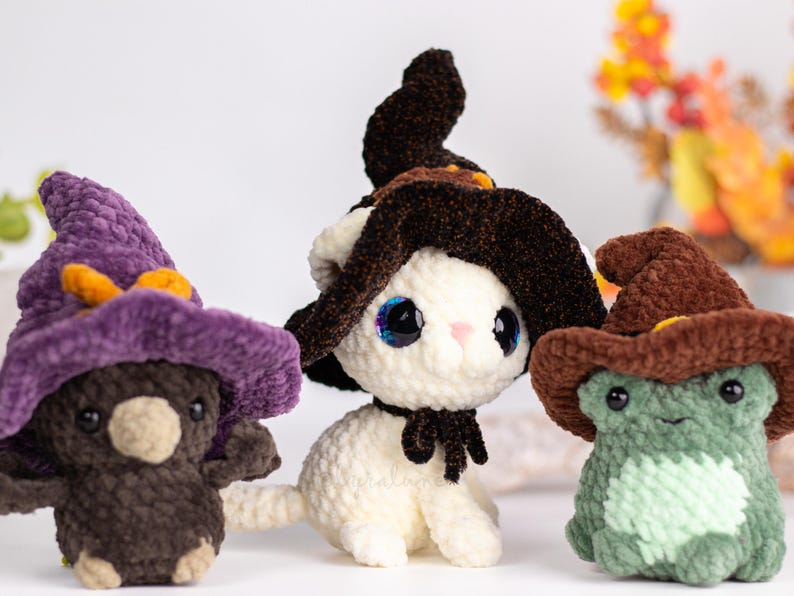 May include: Three handmade crochet plushies: a brown bear, a white cat, and a green frog, each wearing a witch's hat. The bear has a purple hat with orange accents. The cat has a black bow. The frog has a brown hat.