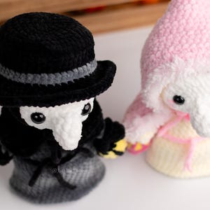 May include: Two crocheted plush toys, one wearing a black hat and a long black coat, the other wearing a pink hooded cloak. Both toys have white faces with black eyes and long white noses.