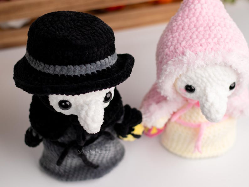 May include: Two crocheted plush toys, one wearing a black hat and a long black coat, the other wearing a pink hooded cloak. Both toys have white faces with black eyes and long white noses.